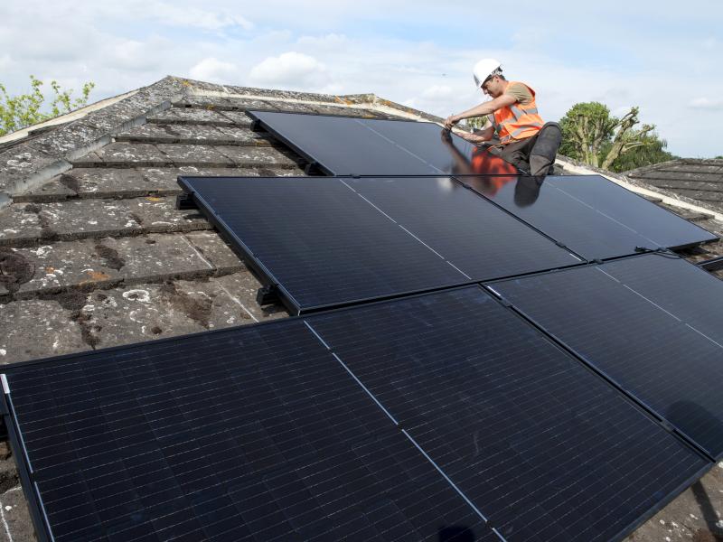 An installer sat on a roof securing solar panels to a tiled roof on a sunny day