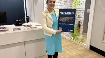 Cllr Qais holding a Here2Help poster. She is wearing a blue dress with a white jacket and standing in the council house reception.