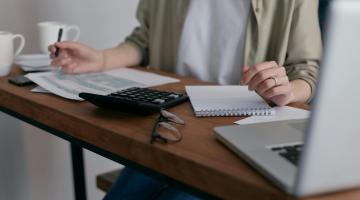 Woman sitting behind a desk with a calculator, notepad, glasses and laptop on the table in front of her. She has a pen in her hand.