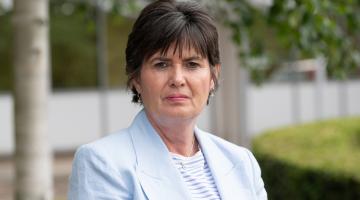 Cllr Karen Grinsell stands outside the council house with greenery around her, she has a neutral expression on her face. 